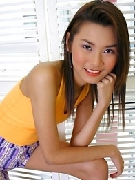 Cat Chonthicha sits on a bed and hugs a pillow. She has on a yellow top and she is looking so sexy as she smiles at the camera.